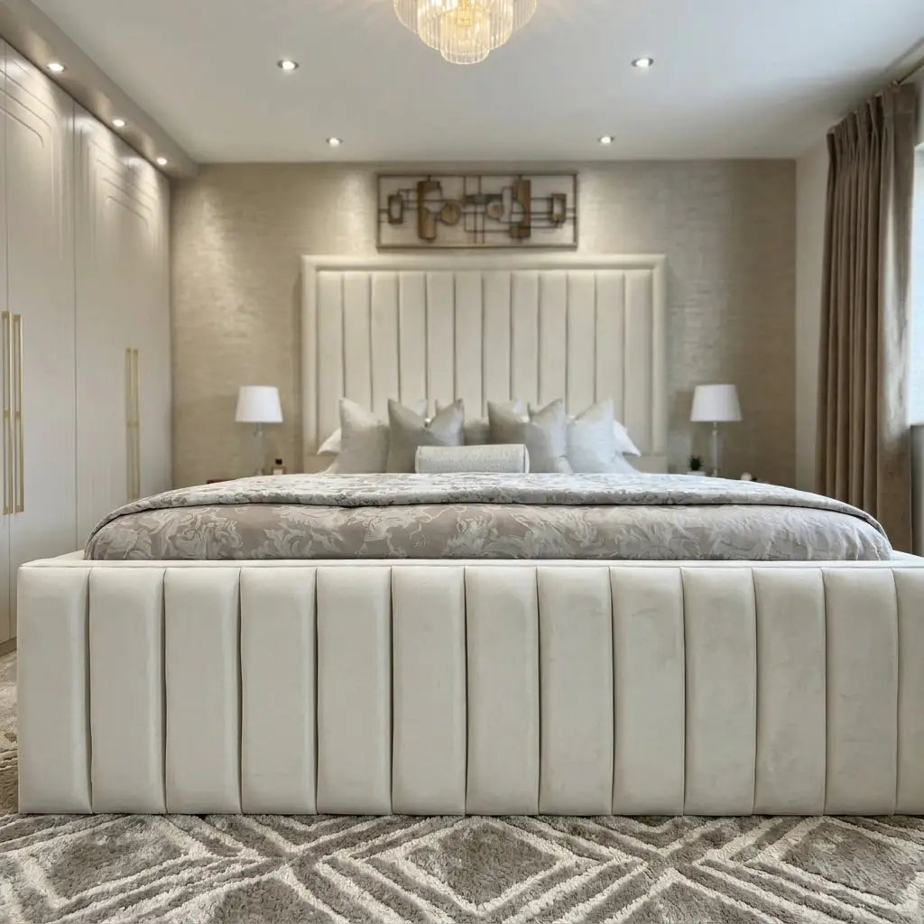 A luxury upholstered bed with a tall channel-tufted headboard and matching footboard in cream velvet, styled in a modern beige-toned bedroom with a geometric rug, bedside tables, and lamps.