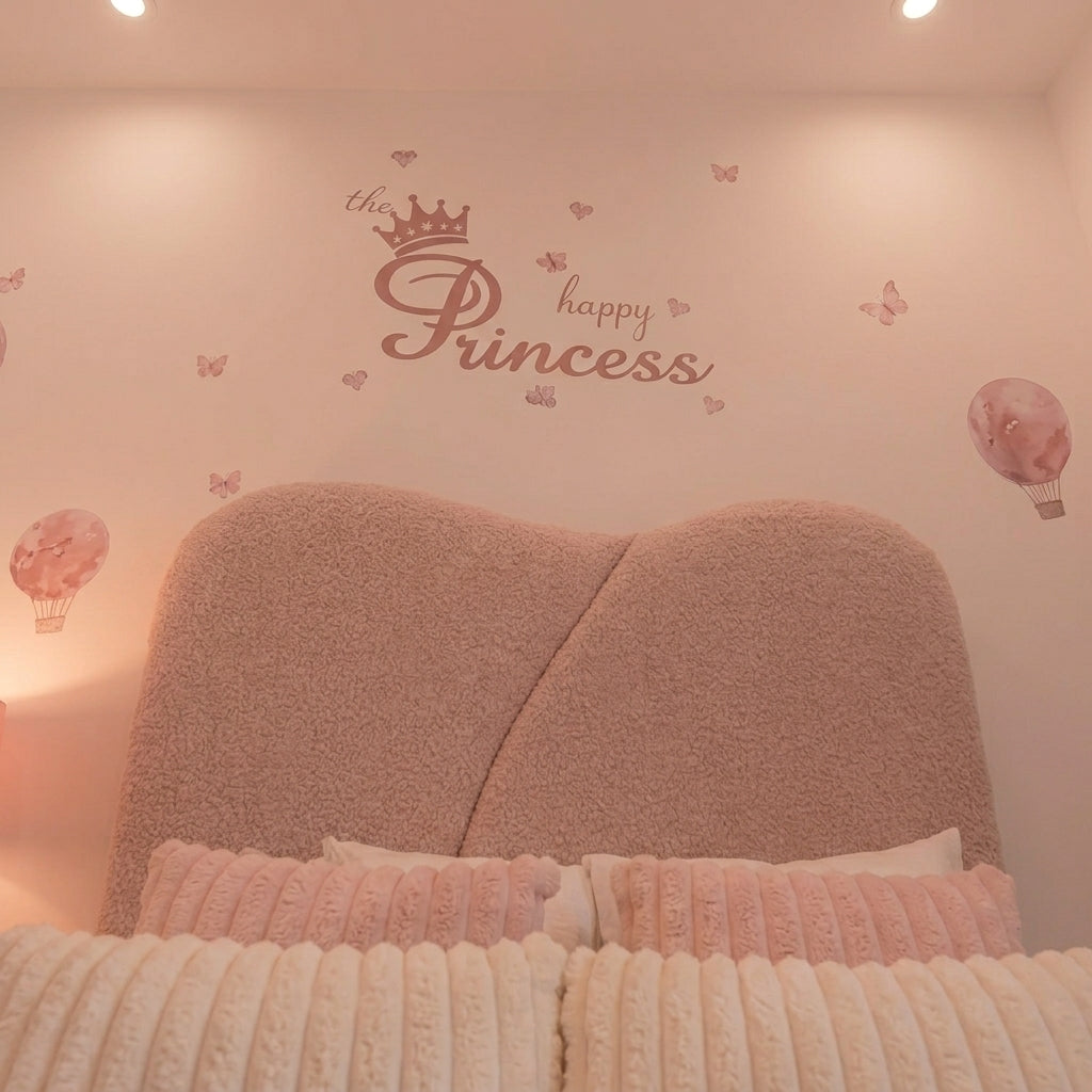 A pink 'the Princess happy' wall decal with a crown and butterflies is displayed above a heart-shaped bed headboard in a cozy, softly lit bedroom.