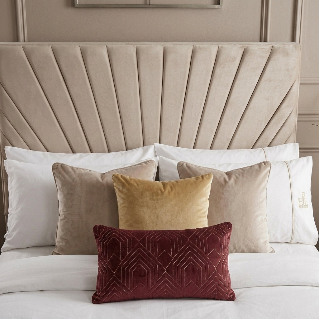 A styled bedroom scene with a large beige upholstered headboard, layered white and neutral pillows, and a prominent burgundy rectangular lumbar pillow with geometric embroidery in the foreground.