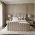 A beige upholstered bed with a vertically channel-tufted headboard and matching footboard, styled in a symmetrical, elegant bedroom with neutral beige walls, matching nightstands, and lamps.