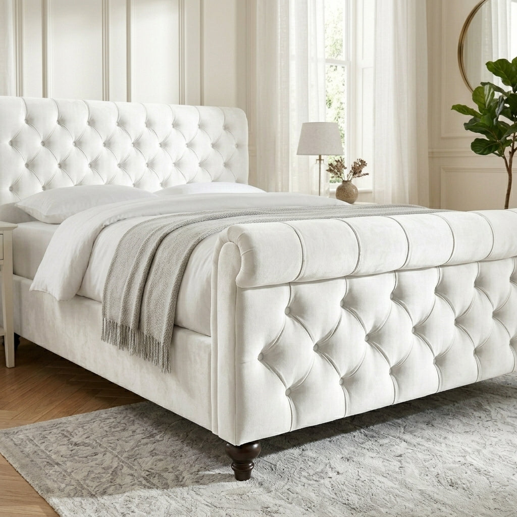 A white tufted velvet bed frame with dark wooden legs, styled with white bedding and a light gray throw blanket in a bright, classic bedroom setting.