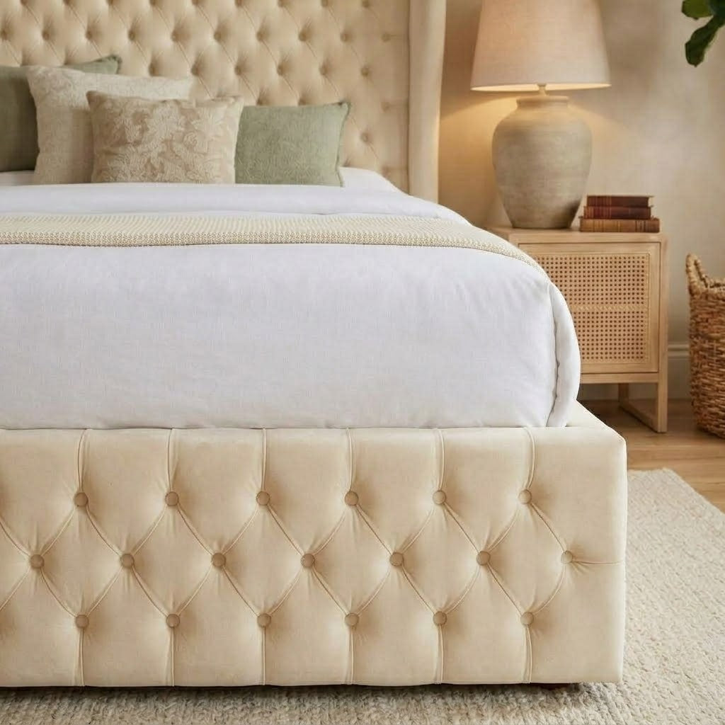 A cream tufted upholstered bed frame with a matching headboard, styled in a warm neutral bedroom setting with white bedding, green accent pillows, a wooden nightstand, and a lamp.