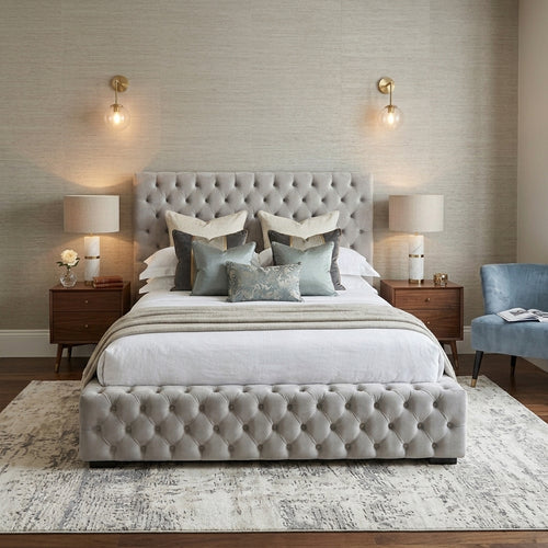 A luxury tufted platform bed in a modern bedroom setting, featuring a light gray upholstered headboard and matching footboard, styled with white bedding, gray and blue accent pillows, wooden nightstands, and wall sconces.