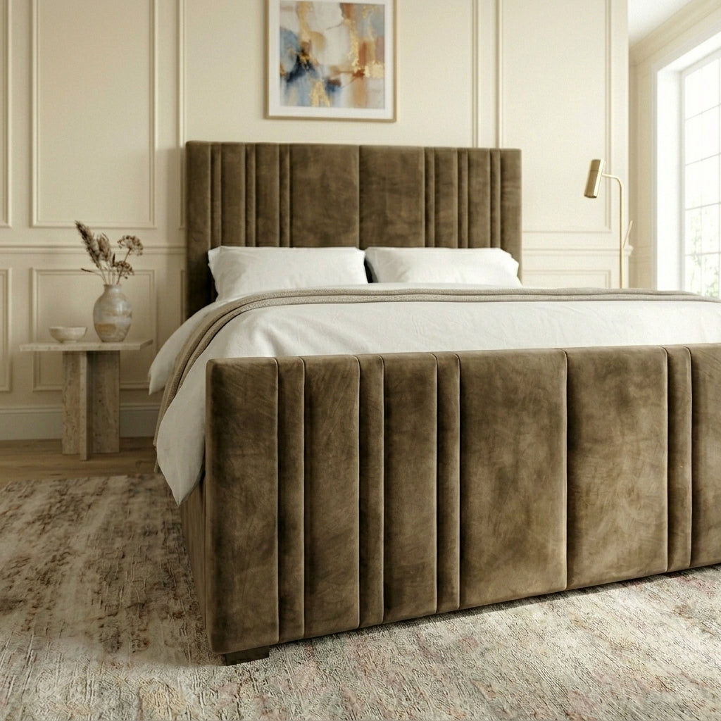 A bed with a tall, pleated upholstered headboard and matching footboard in a warm taupe-brown velvet fabric, styled in a bright, modern bedroom with white bedding and a textured rug.