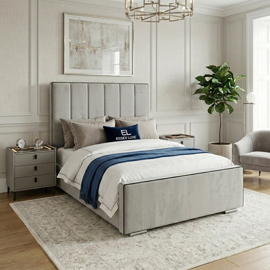 A modern upholstered bed in a styled bedroom setting. The bed features a light gray fabric frame with a vertically channel-tufted headboard and matching footboard, dressed in white bedding with a navy blue throw blanket and a navy blue accent pillow that reads 'ESSEX LINE'.