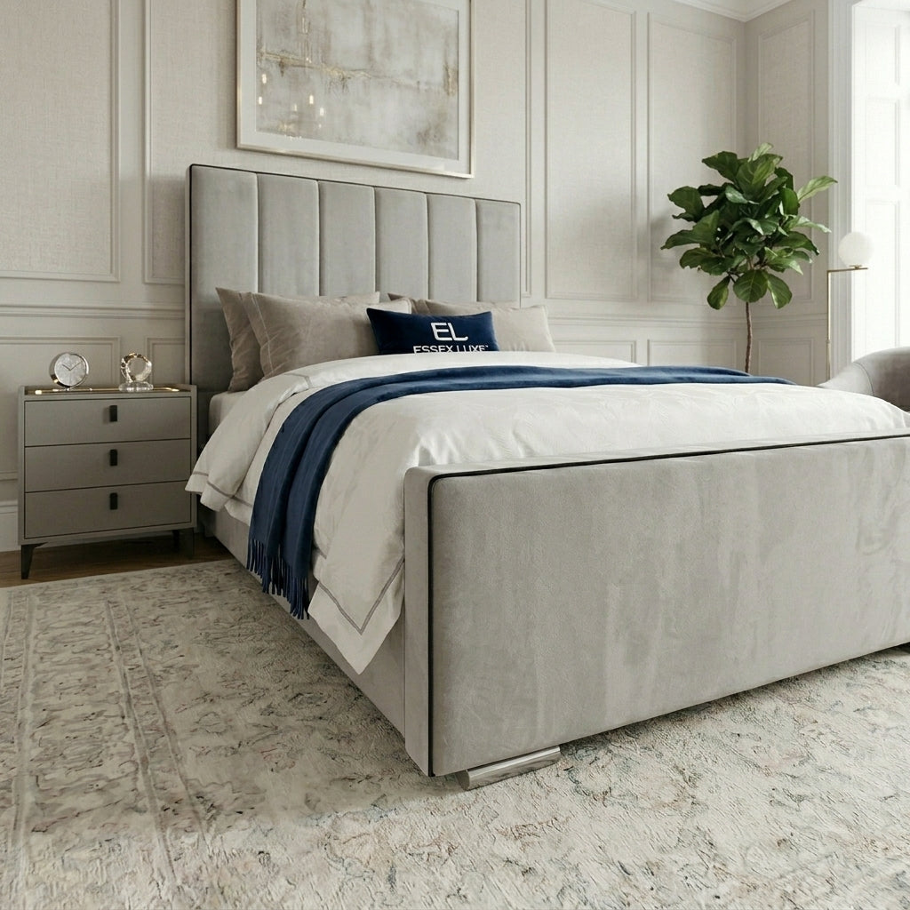 A modern upholstered bed in light gray fabric with a tall segmented headboard and matching footboard, styled in a bright bedroom with white bedding, a navy throw blanket, gray pillows, a nightstand, rug, and potted plant.